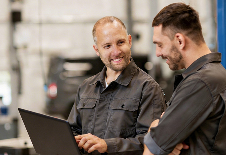 Zwei Männer in Arbeitskleidung sprechen in einer Industriehalle vor einem Laptop miteinander.