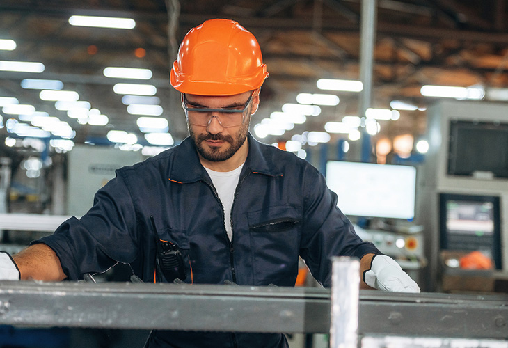 Mann mit orangefarbenem Schutzhelm und dunkelblauer Arbeitsjacke arbeitet mit Stahl in einer Industriehalle. Mann mit orangefarbenem Schutzhelm und dunkelblauer Arbeitsjacke arbeitet mit Stahl in einer Industriehalle.