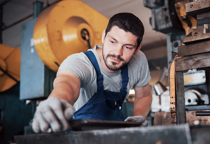 Junger Mann in blauer Arbeitskleidung und Schutzhandschuhen arbeitet konzentriert an einem Metallteil in einer hellen Werkstatt. Junger Mann in blauer Arbeitskleidung und Schutzhandschuhen arbeitet konzentriert an einem Metallteil in einer hellen Werkstatt.