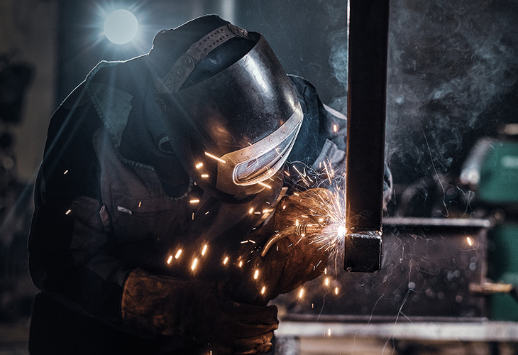 Schweißer mit Schutzvisier und schwarzen Handschuhen schweißt wo zahlreiche hell-orange leuchtende Funken sprühen.